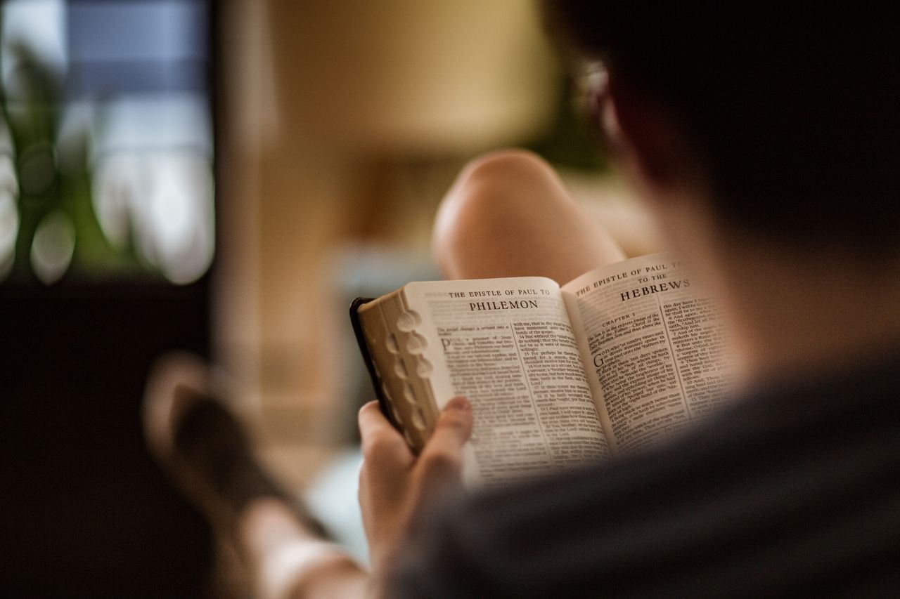 Një burrë i ulur në një kolltuk, studion Biblën e Shenjtë me faqen të hapur te letra e Palit drejtuar Filemonit
